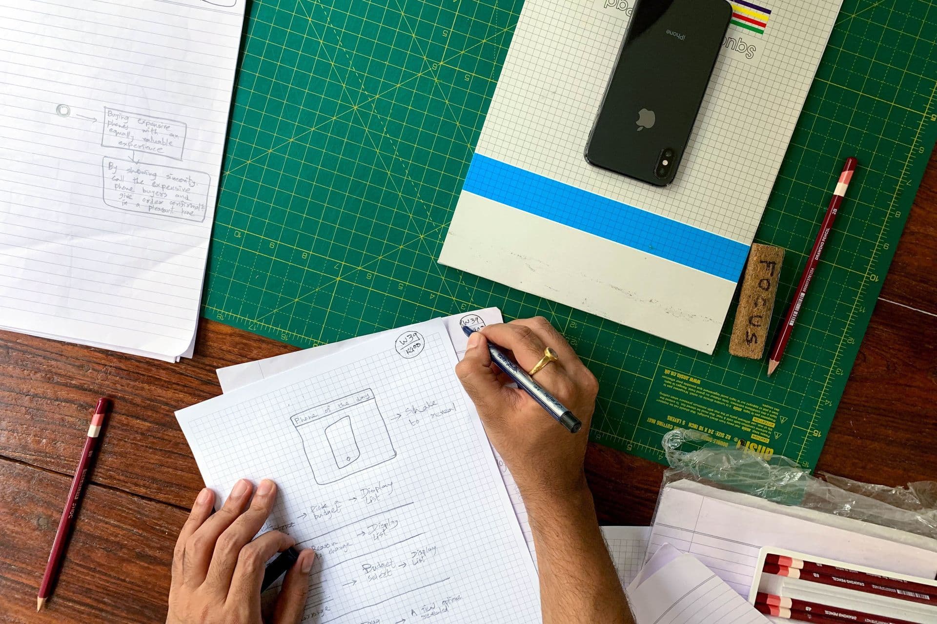 Hands sketching a mobile app wireframe on graph paper beside a phone and pencils