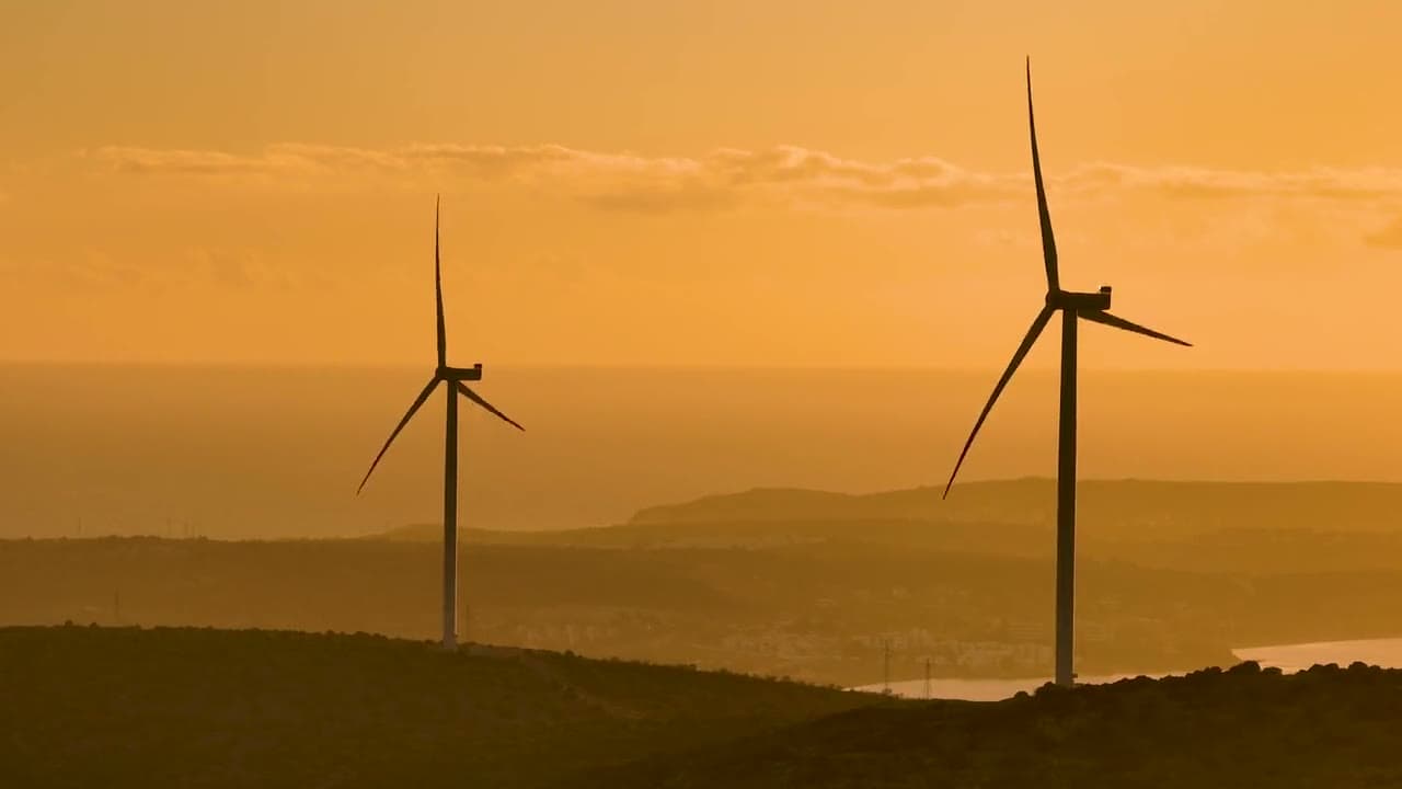 Two wind turbines silhouetted on a hill against an orange sunset sky and sea