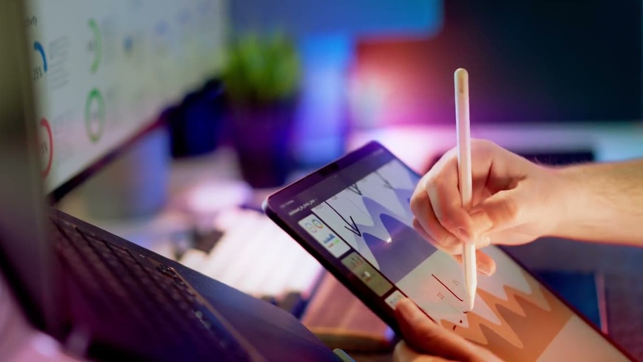 Hand with stylus on a tablet showing charts, monitor with dashboard graphs behind