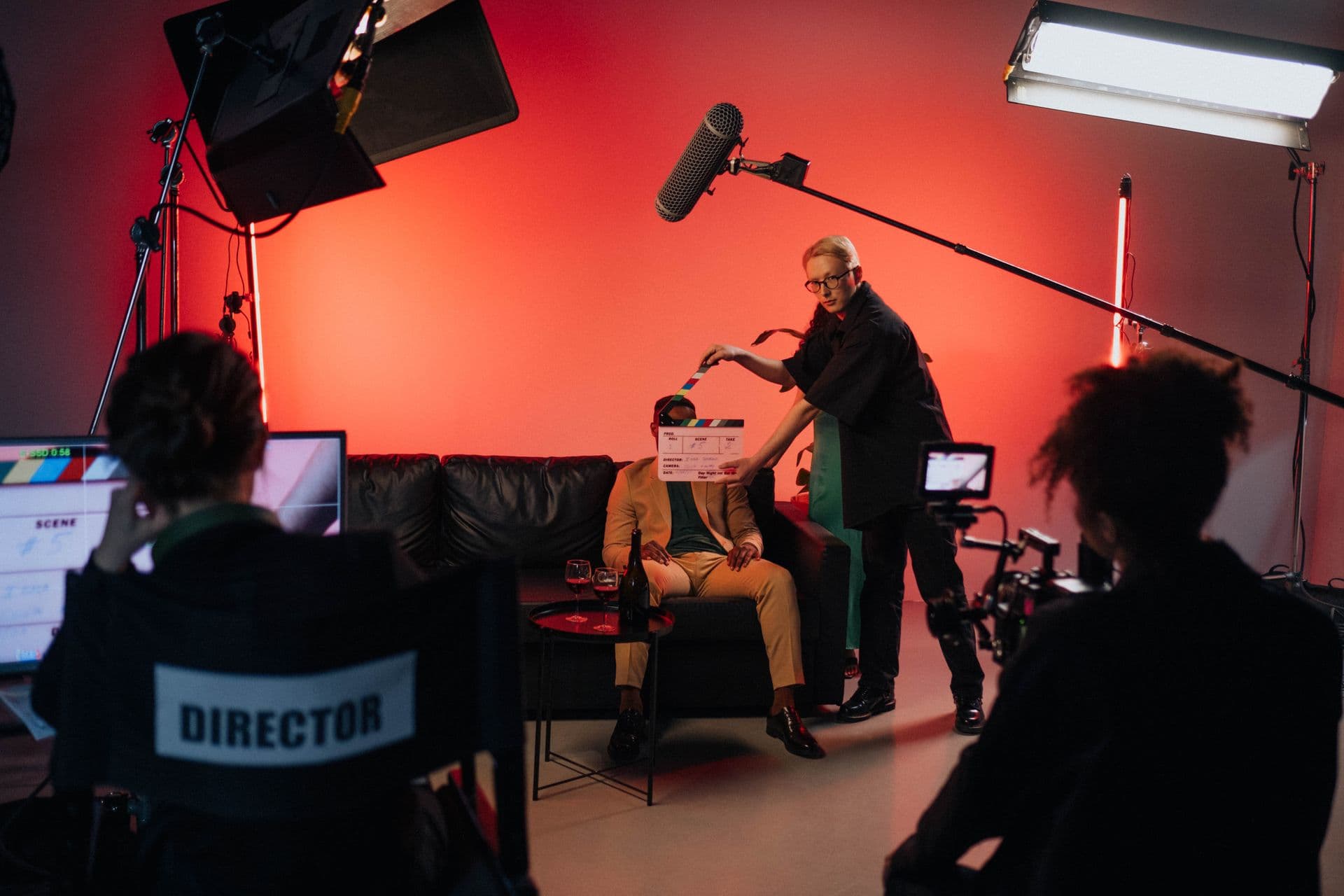Film crew on a red-lit studio set with a clapperboard, boom mic and director's chair