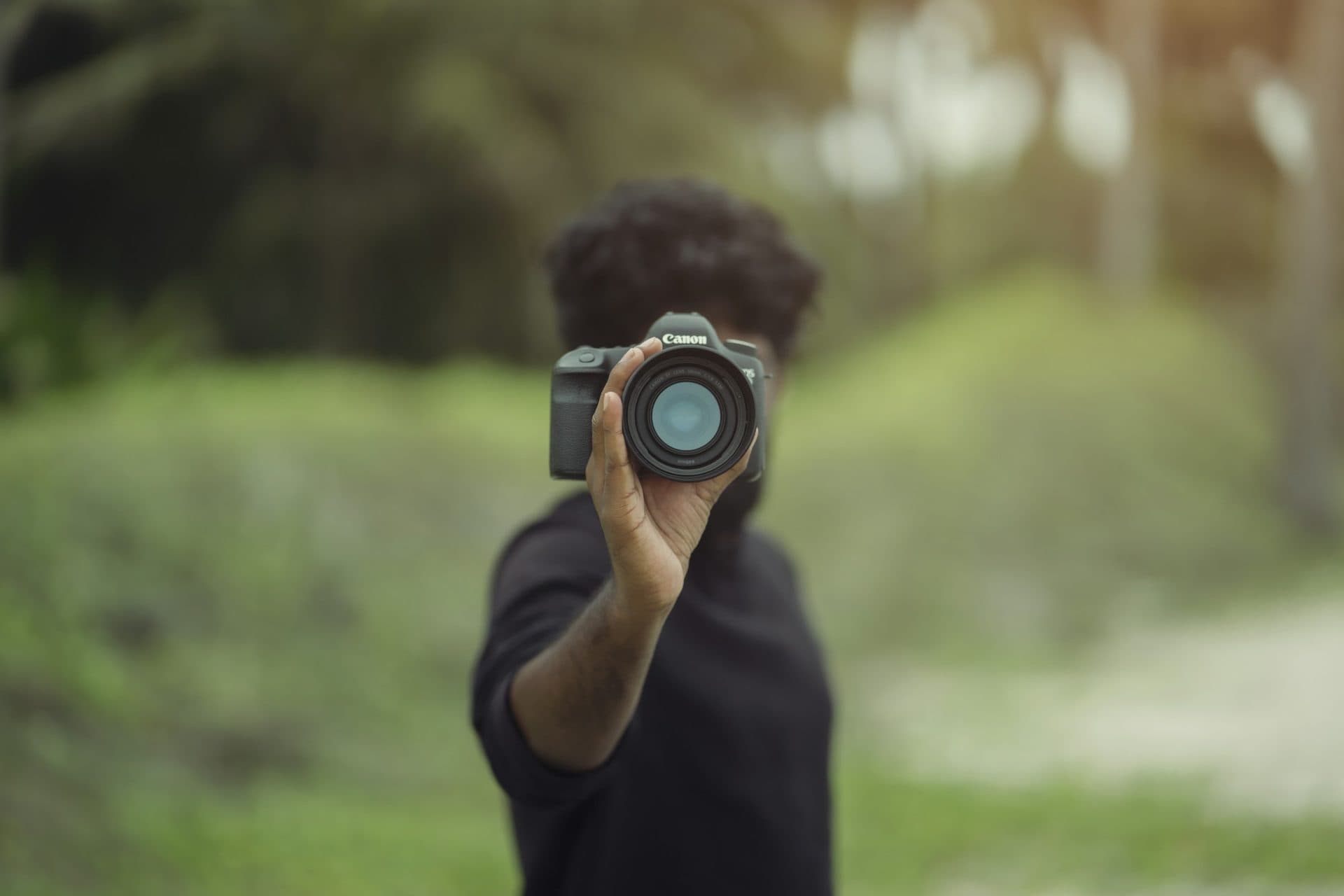 Man holds a camera toward the viewer against a soft green, blurred background