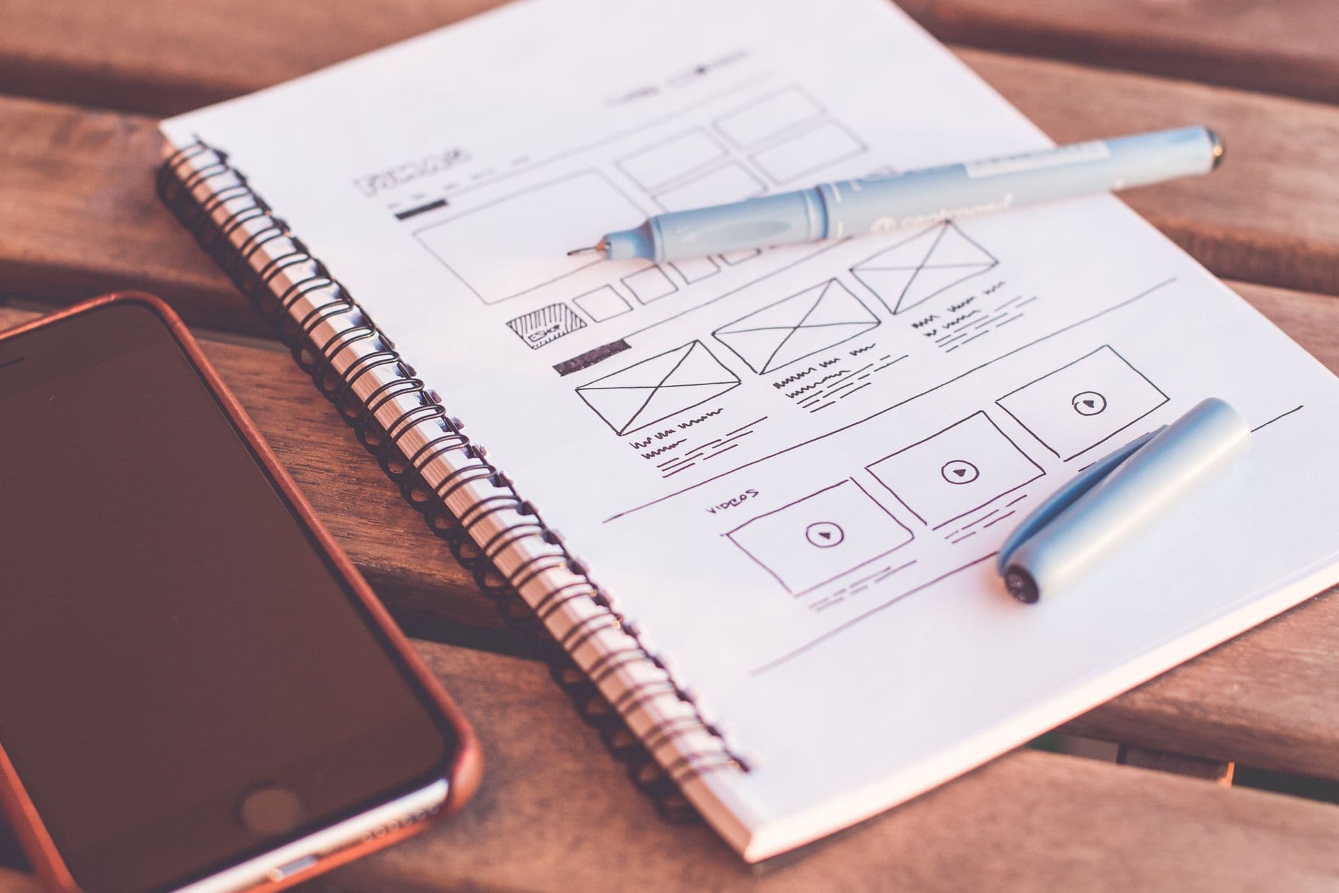 Website wireframe sketches in a spiral notebook next to a smartphone and pens on a desk