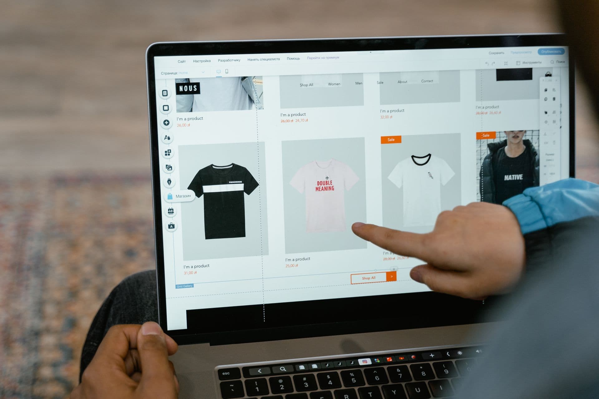 Hand pointing at T-shirt products on an online store page shown on a laptop