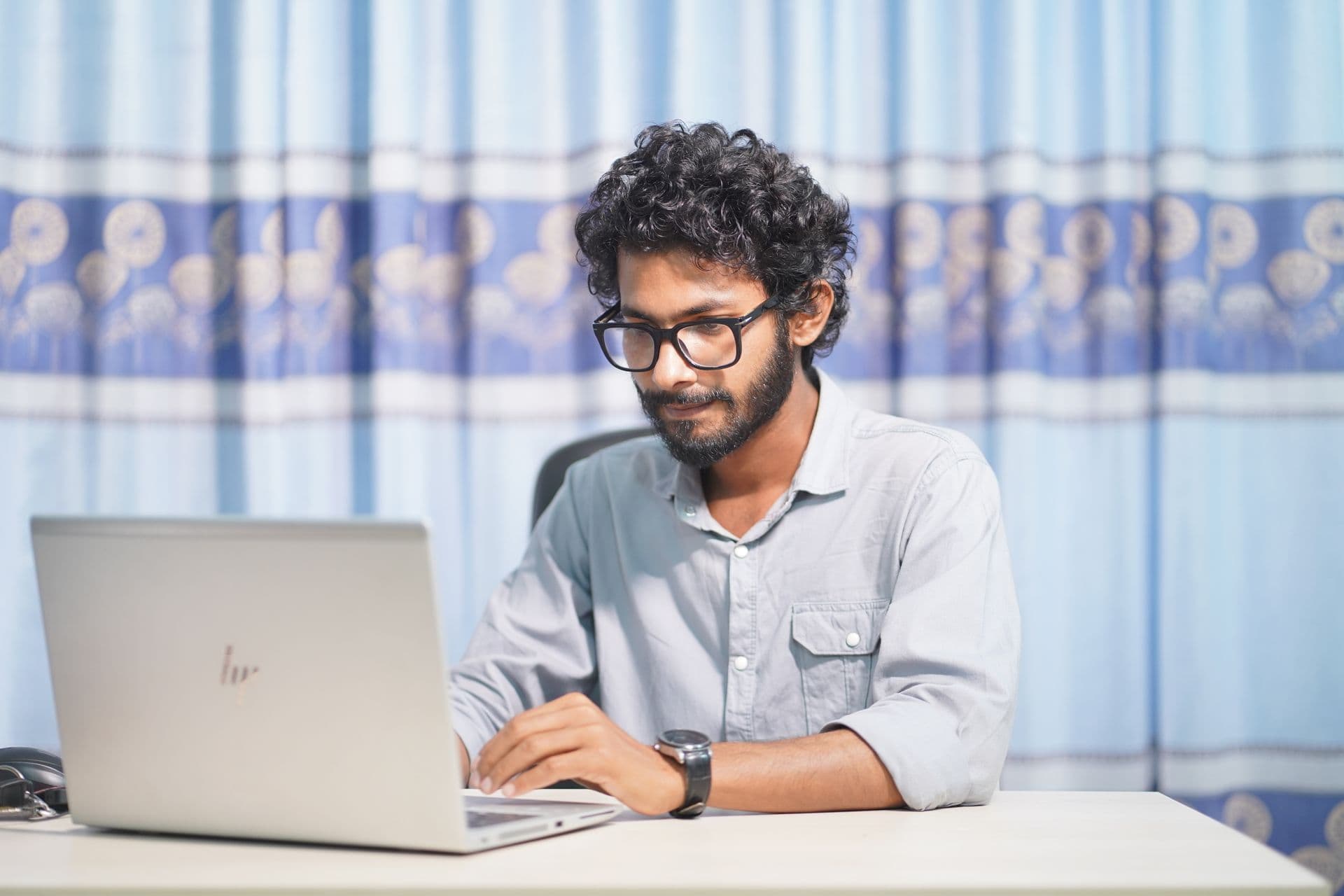 Young man with glasses and curly hair typing on a laptop at a white desk