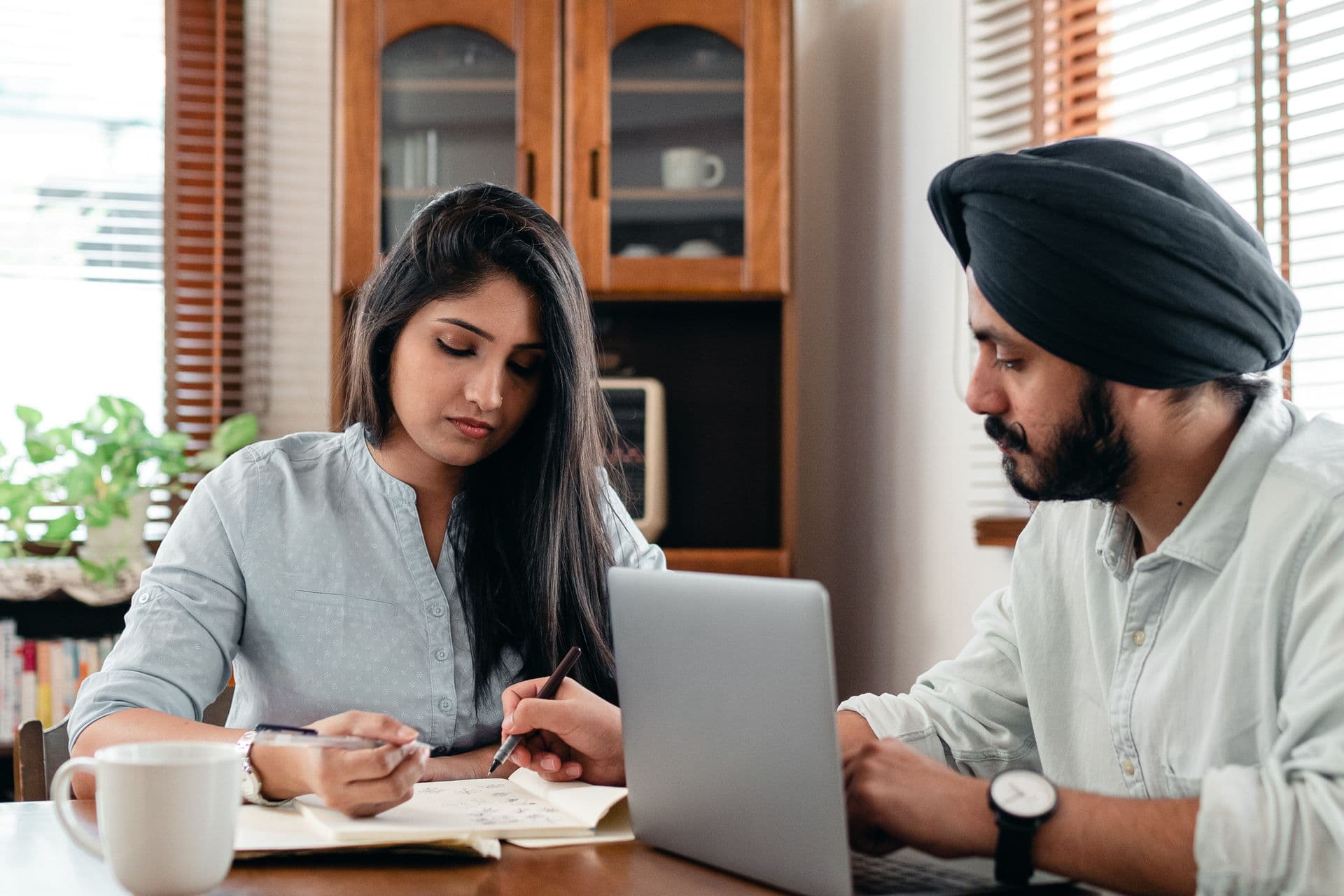 Man in a turban types on a laptop while a woman takes notes beside him