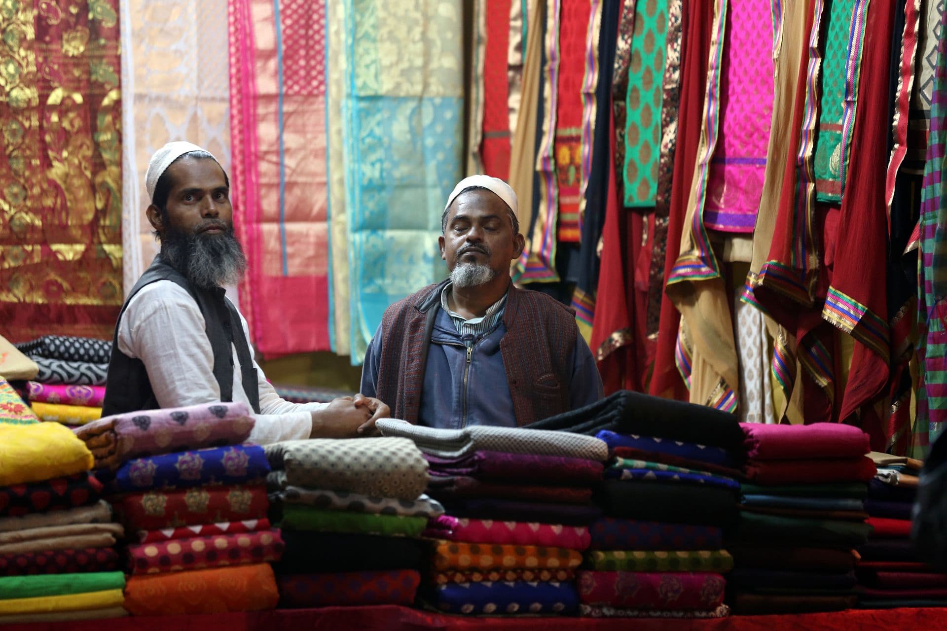 Two men seated behind stacks of colourful folded fabric with sarees hanging behind them