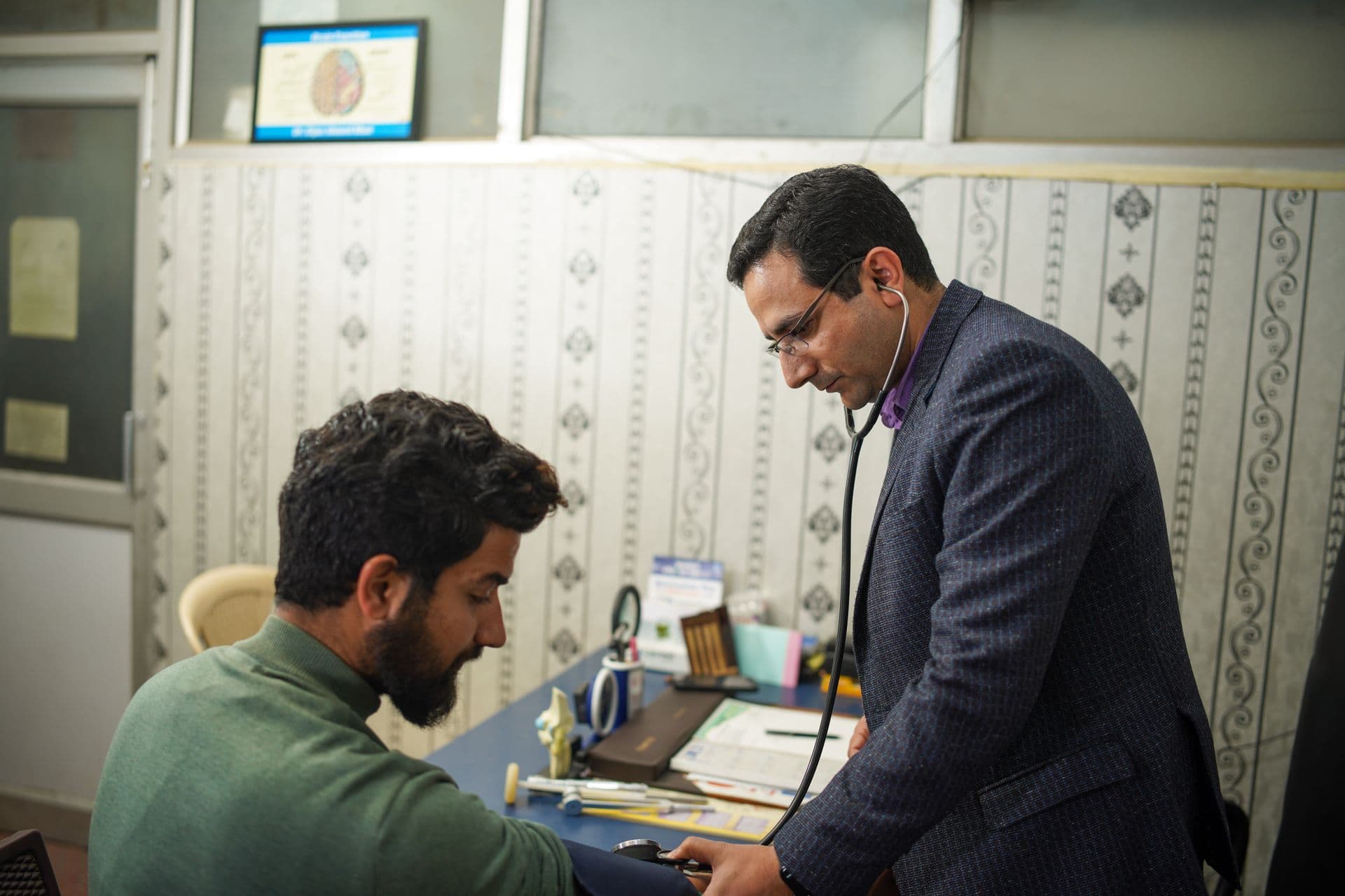 Doctor in a blazer checking a seated patient's blood pressure with a stethoscope in a clinic