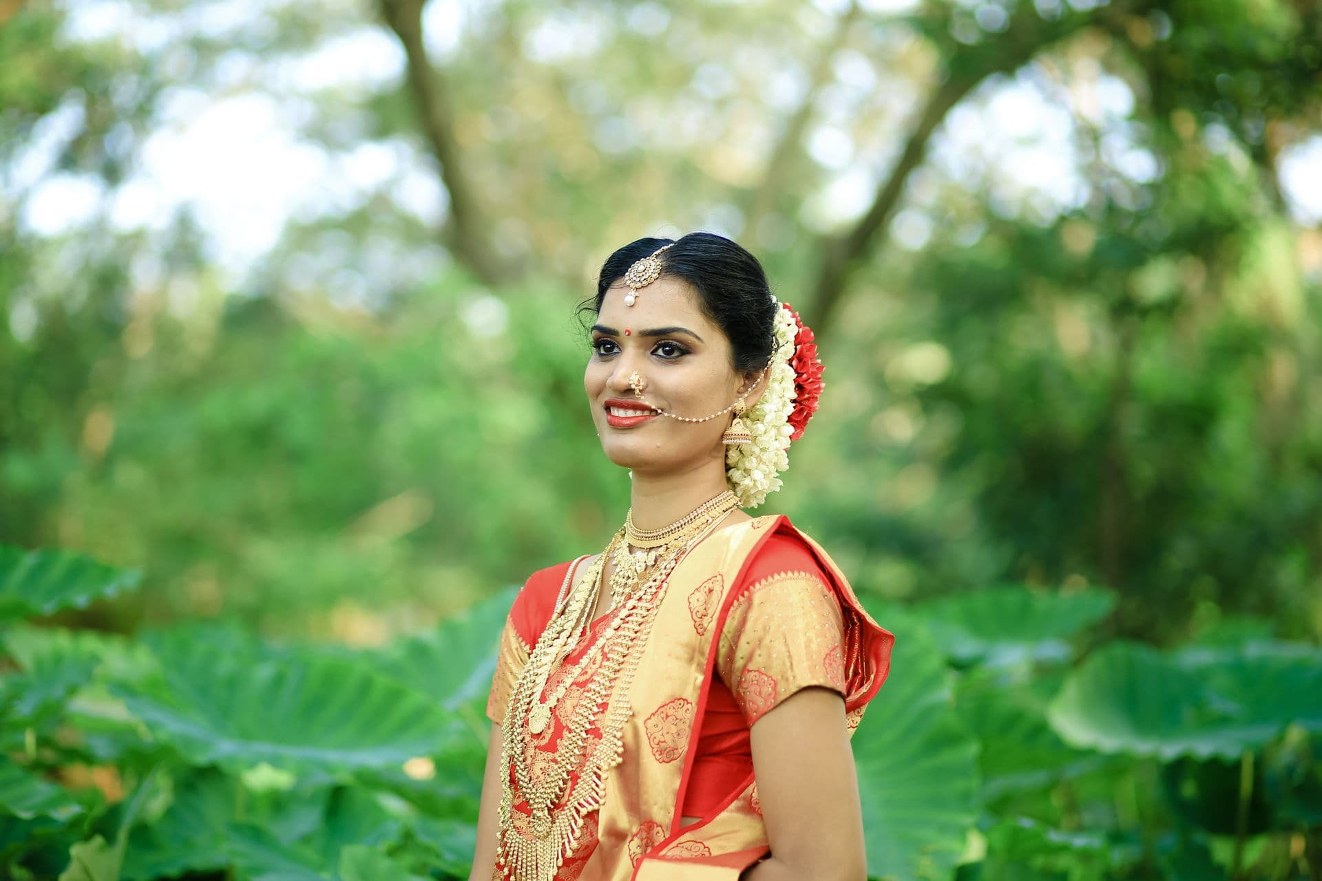 Smiling bride in a red and gold saree wearing layered gold necklaces and jasmine flowers