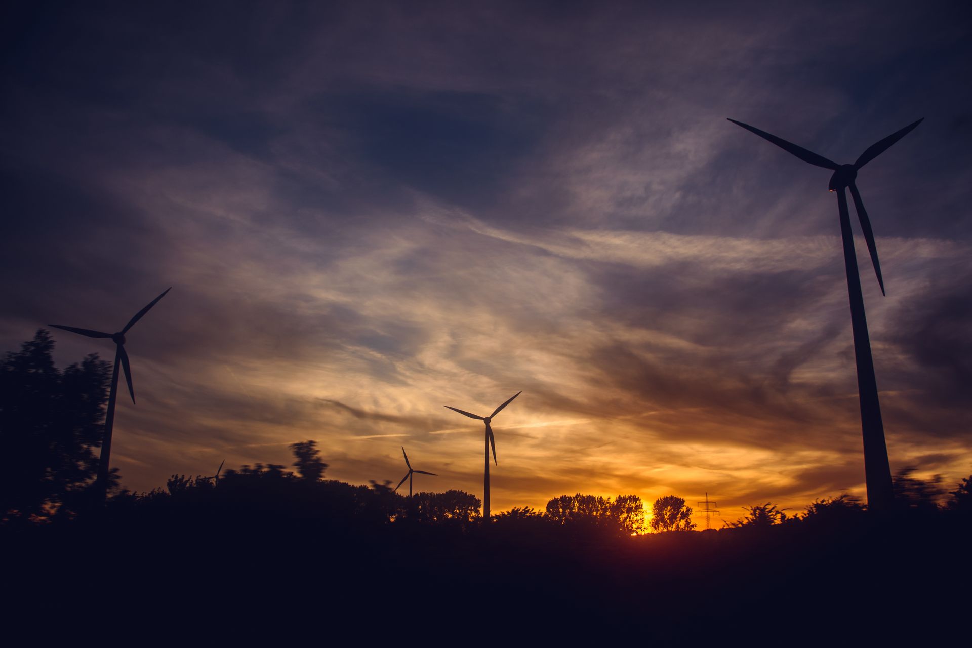 Silhouetted wind turbines against a dramatic orange sunset sky above a line of trees