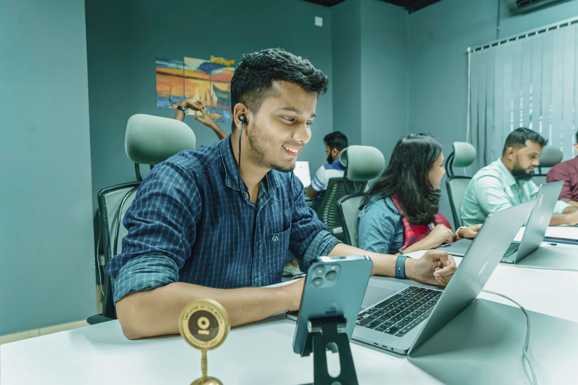 Smiling young man works on a laptop with a phone on a stand beside him