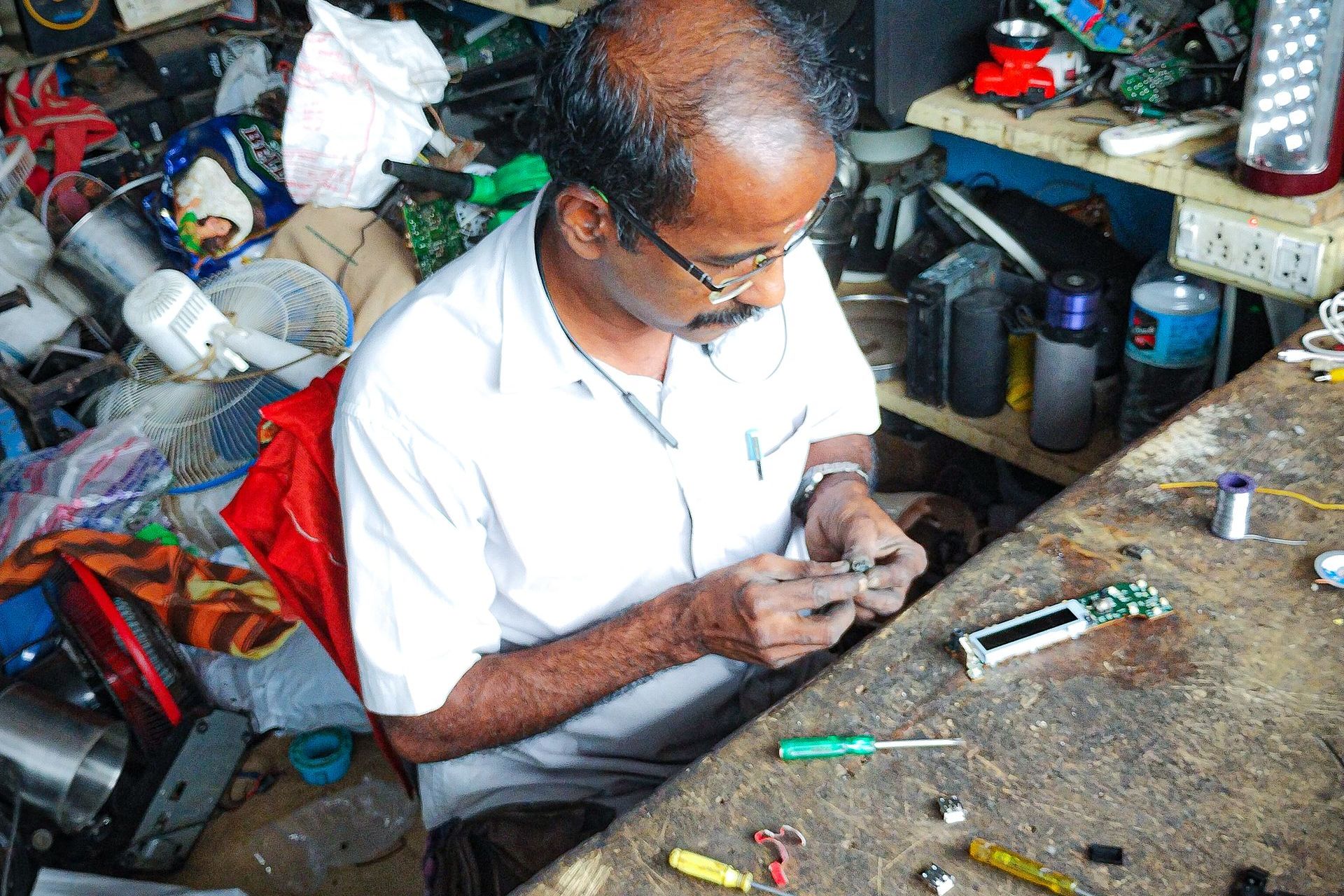 Technician in glasses repairing a small electronic part at a cluttered workbench